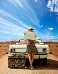 Femme assise sur le capot de sa voiture en train de regarder une carte routière avec sa valise posée par terre