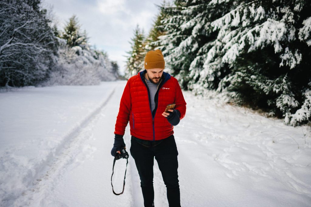 homme portant une doudoune rouge un pantalon noir et un bonnet tenant un appareil photo et regardant son portable sur un chemin enneigé